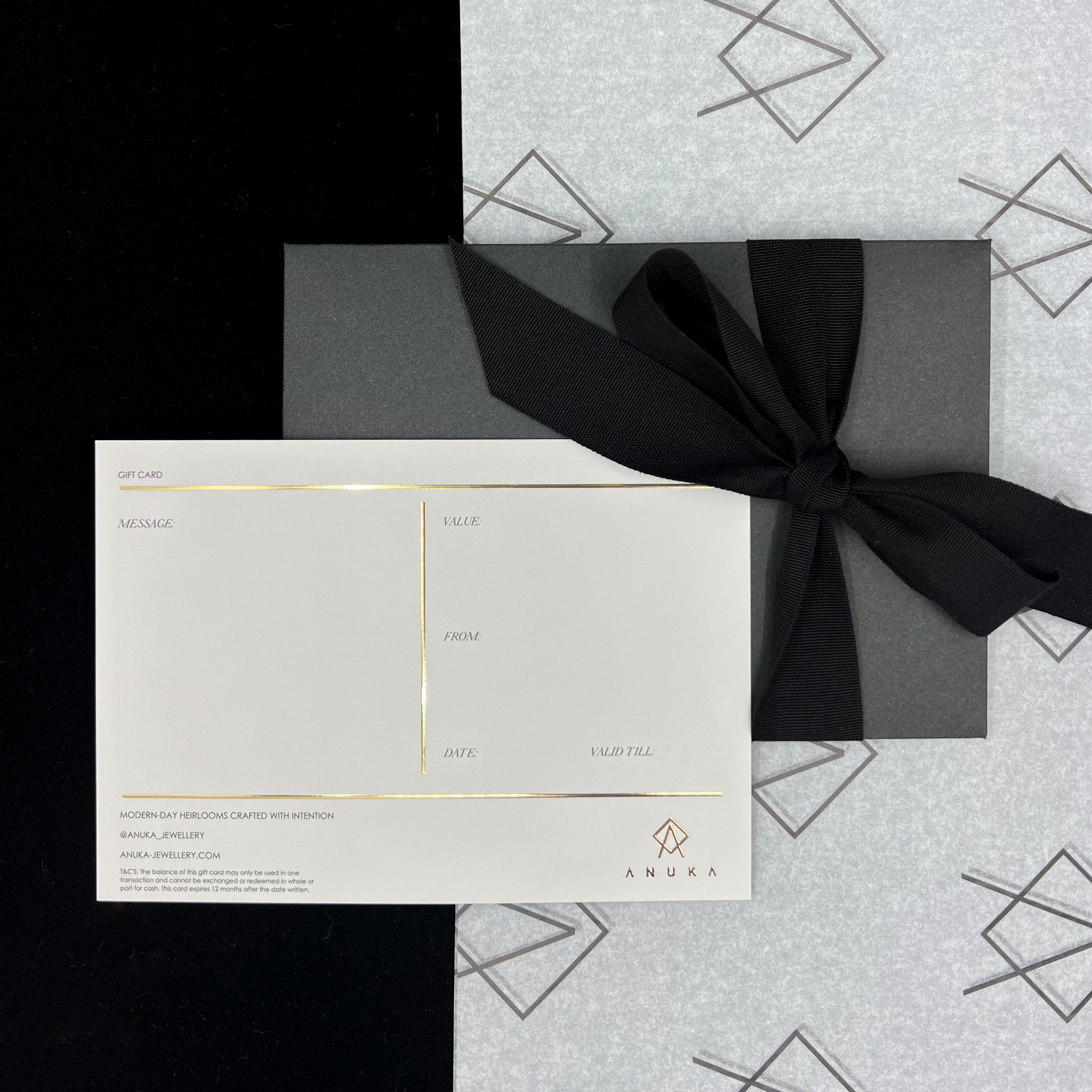A gift voucher from ANUKA Jewellery, featuring a card wrapped in a black envelope with a decorative silver ribbon, presented on a background with geometric pattern.